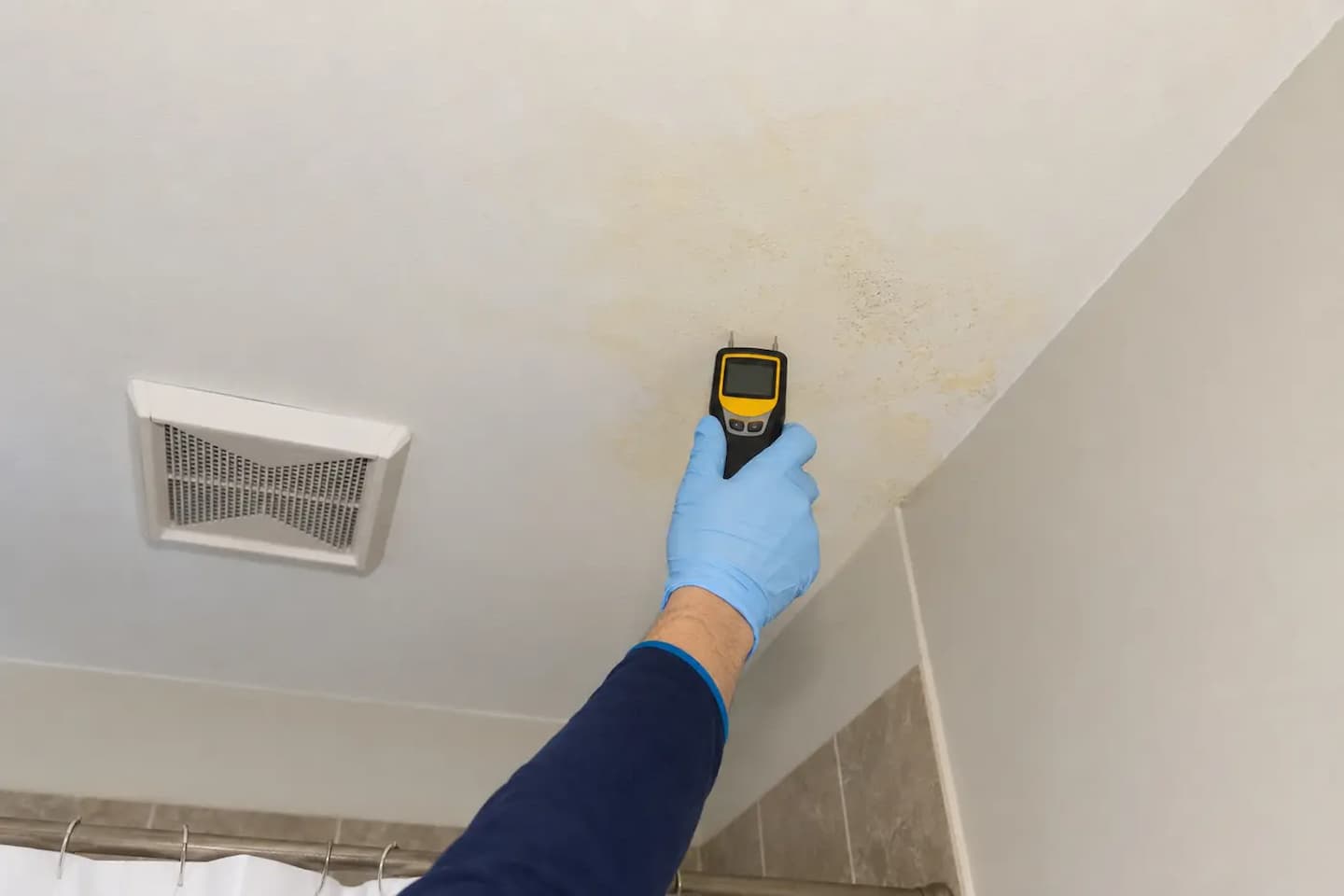 Gloved hand using a moisture meter near yellow discoloration on a bathroom ceiling